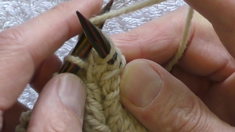 The left hand needle passes behind the right hand needle from bottom left to top right through the stitch on the right hand needle. It is being swung back and to the left to catch a loop of the working yarn that is held above and to the right of the needle tips by the index finger of the right hand.