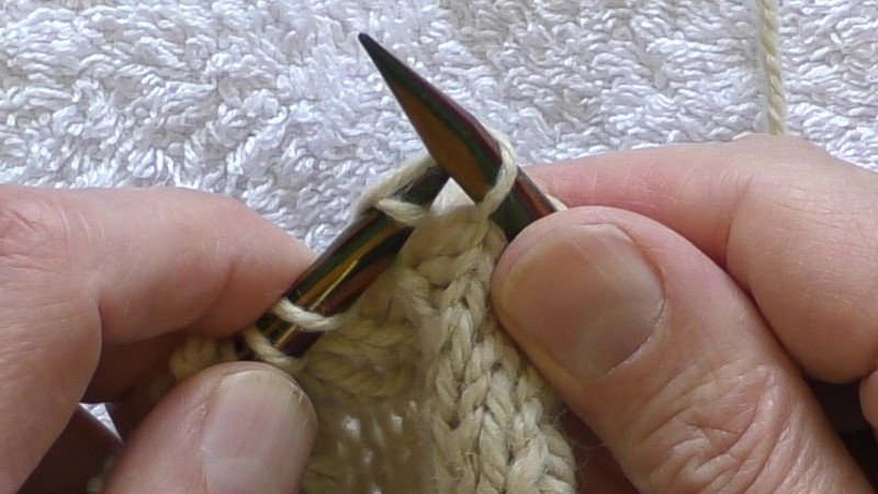 The tip of the left hand needle with a loop of working yarn passing to the front of it is being pulled forwards through the centre of the stitch on the right hand needle.