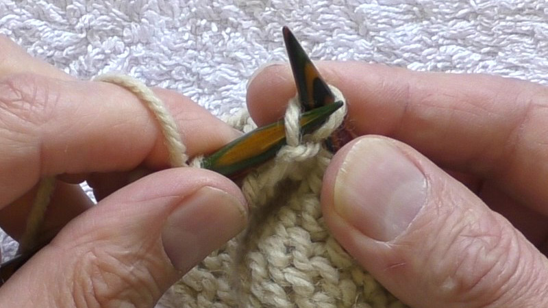 The purl side of the fabric is facing the viewer. Stitches already worked are on the left hand needle. The working yarn is in the left hand. The left hand needle has been placed through the centre of the first stitch on the right hand needle, passing from back right to front left. The left leg (that was at the back of the right needle) lies across the front of the left needle, and the right leg (that is on the front of the right needle lies behind the left needle. The tip of the left needle is in front of the right needle.