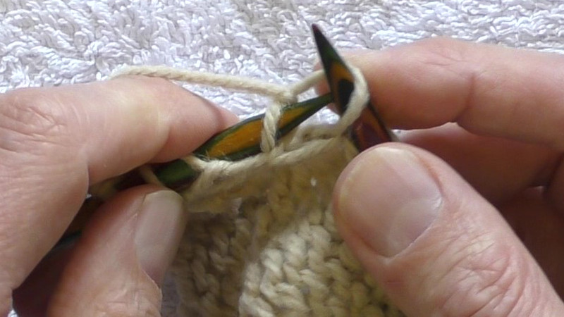 The working yarn has been placed around the left hand needle from left to right so that the working yarn passes below the left hand needle with the free end behind the left hand needle. The swing continues to take a loop of the working yarn to the back of the fabric through the centre of the old stitch on the right hand needle.