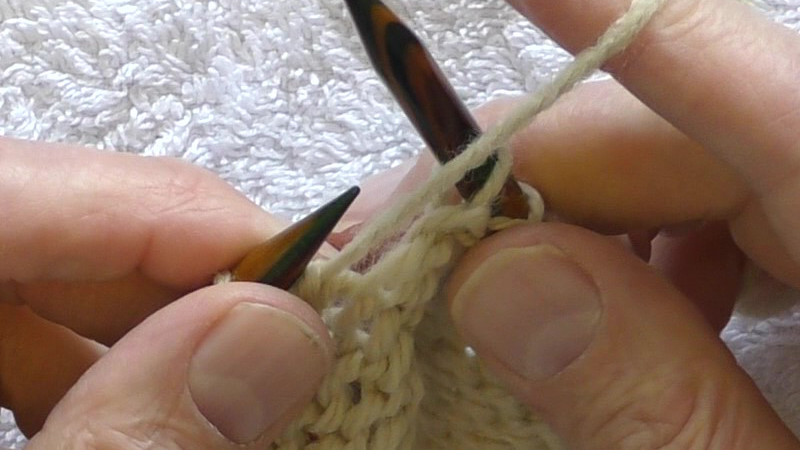 The new stitches of the current row are on the left hand needle. Stitches from the previous row are on the right hand needle. The yarn is being held to the front of the fabric and passes from the front over the index finger of the right hand.