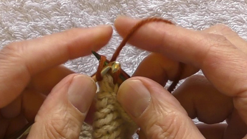 Right hand needle being placed through the loop on a crochet hook so that the stitch is "western mounted" with the running thread from the previous stitch to the front of the needle and the supply of working yarn behind the needle.