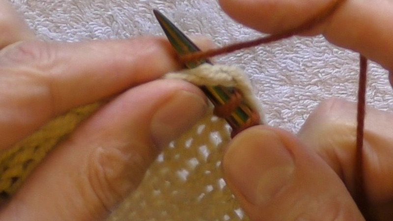 Working yarn being wrapped around the needle for a knit stitch. The needle has been pushed through the centre of the stitch immediately below the cast-off edge, with the cast-off chain lying on the top of the needle.