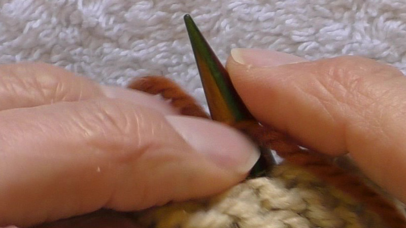 Tip of needle poking through the fabric to the private (wrong) side. The working yarn lies across the near (top) side of the needle, with the free end to the right of the picture.