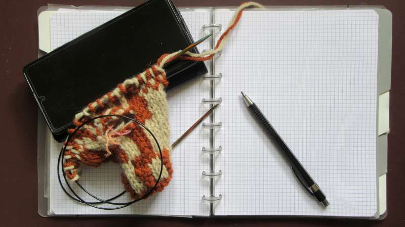 Notebook under knitted work in progress, mobile phone and a pencil.
