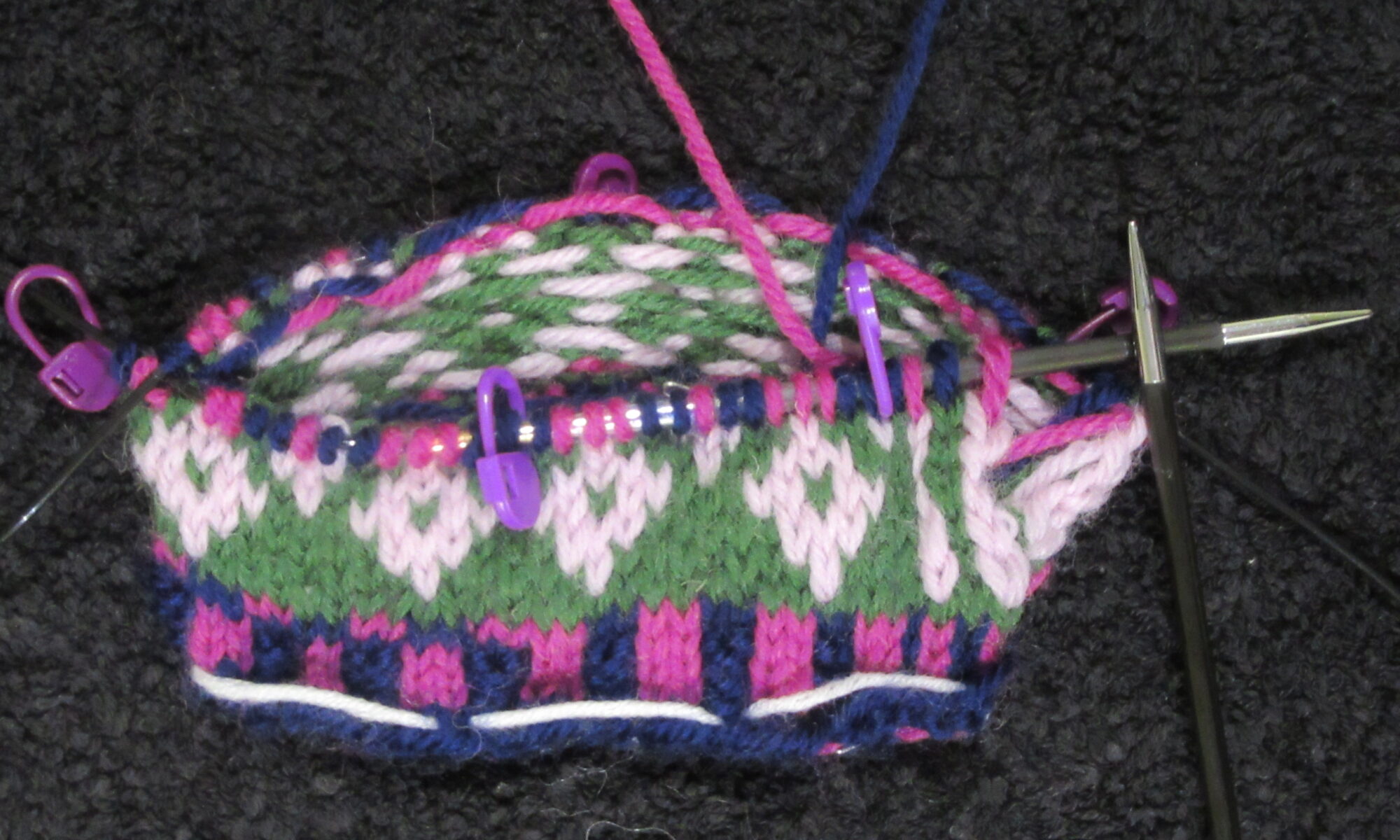 Stranded knitting being worked in the round on circular needles using the magic loop. Three rounds of vertical stripe rib are followed by eight rounds of colour work. Stitch markers show the start of pattern repeats.