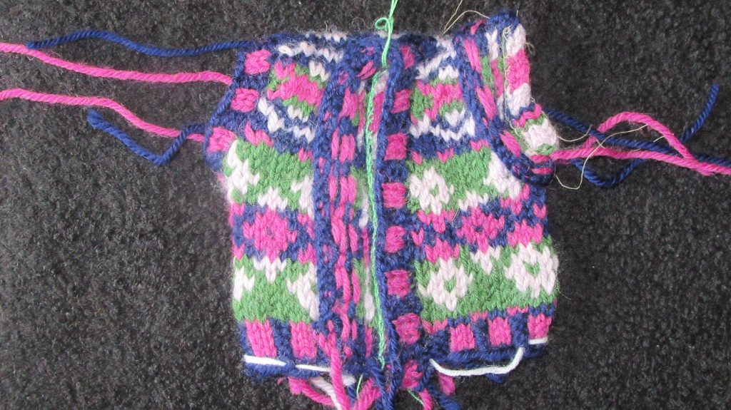 The waistcoat of the Introduction to stranded knitting project after the button- and arm- bands have been added. One of the armholes is shown normally, the other with the band folded back to show the private side. One side of the button band has been folded back to show the private side.