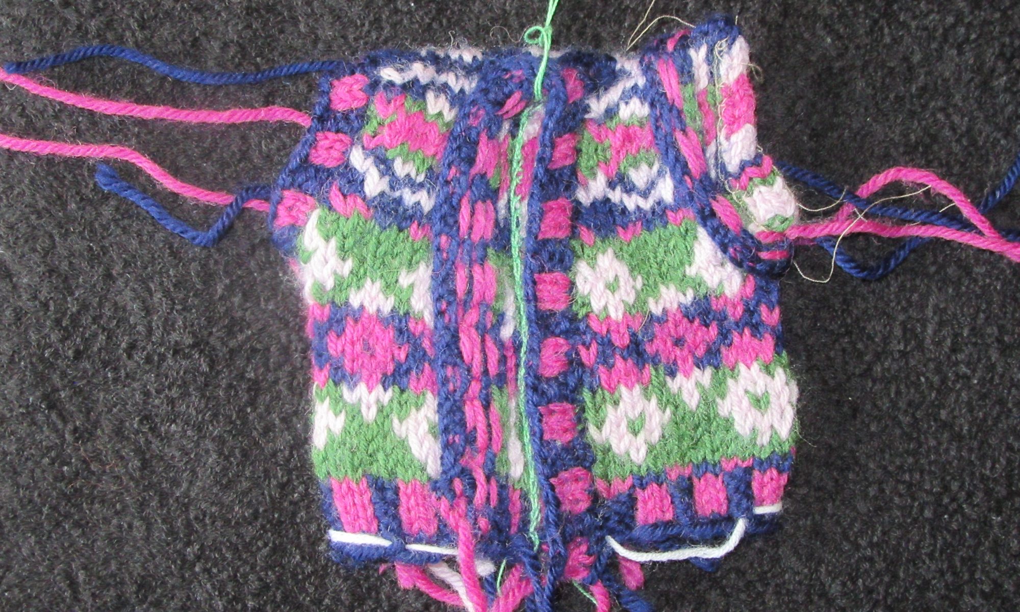The waistcoat of the Introduction to stranded knitting project after the button- and arm- bands have been added. One of the armholes is shown normally, the other with the band folded back to show the private side. One side of the button band has been folded back to show the private side.