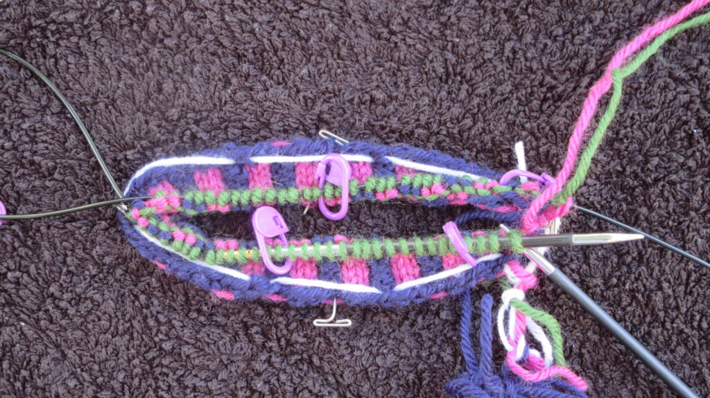 Stranded knitting being worked on circular needles using a magic loop. There are three rounds of vertical stripe rib, one round of stranded colour work and one round of a new background colour. Stitch markers show the starts of pattern repeats.